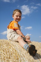 Little boy sitting on a straw bale under a cloudy blue sky