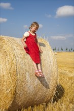 Little girl sliding from a straw bale under a cloudy blue sky in a harvested cornfield