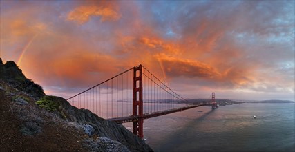 Panoramic view of the Golden Gate Bridge with a rainbow at sunset and orange-glowing storm clouds
