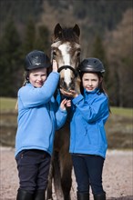 Two girls wearing riding helmets standing beside a pony
