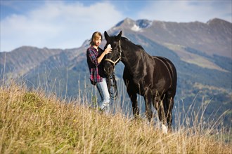A young woman and a black Hanoverian horse standing on a mountain meadow in autumn