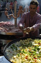 Cook adds pieces of onion to a large pan containing potato stew in front of giant barbecue at the annual All Saints Market in Cocentaina