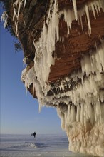 Icicles hanging from red sandstone cliff and people walking on frozen Lake Superior