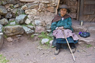 Mature Quechua Indian woman wearing a hat sitting on a stone in front of a doorway