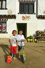 Two boys standing in front of a traditional house with a phallic representation and other symbols of good luck and for the protection against evil spirits