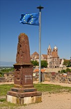 Obelisk and the European flag on Eckartsberg hill in front of the historic centre of Breisach with St. Stephansmunster Cathedral