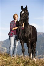 A young woman and a black Hanoverian horse standing on a mountain meadow in autumn