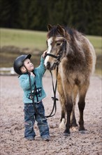 Young child wearing a riding helmet stroking a pony
