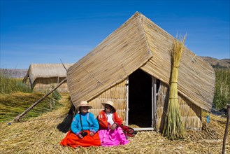 Two women of the Uro Indians wearing traditional dress sitting in front of a reed hut