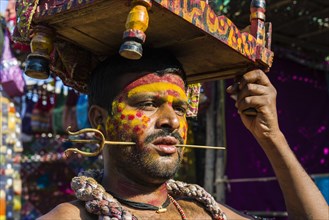 A man with a small brass trident pierced through his cheek is collecting money for religious purposes at the weekly flea market