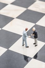 Two figurines of businessmen standing on a chessboard shaking hands