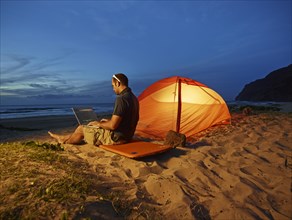 Man with a laptop sitting in front of a tent on a sandy beach at dusk