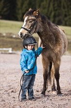 Young child wearing a riding helmet standing beside a pony