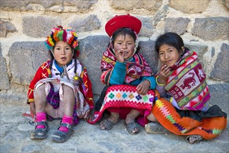 Three children in traditional dress of the Quechua Indians sit in front of a wall