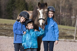 Three children wearing riding helmets standing beside a pony