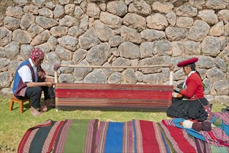 Two Quechua Indians in traditional dress squatting on the floor working on the stretcher of loom