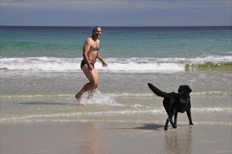 Man in swimming trunks playing with a dog on the beach of Rispond Bay