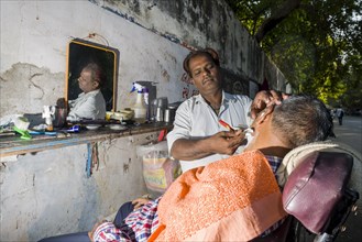 A barber is shaving a customer at his open-air shop
