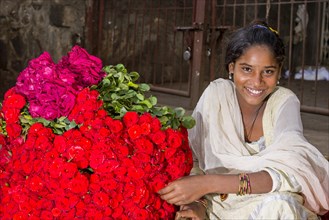A smiling young woman in a white sari is selling red roses at the weekly market