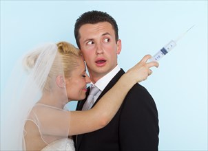 Groom looking puzzled over his shoulder at a syringe in the hand of his smiling bride
