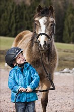 Young child wearing a riding helmet standing beside a pony