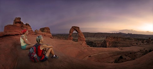 Hikers sitting at Delicate Arch natural stone arch at dusk