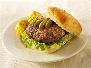 Grilled beef patty on a ciabatta bun with pickles and grilled corn on a plate