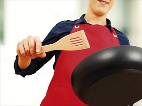 Young man wearing an apron holding a pan and a spatula