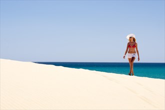 Woman wearing a hat and a bikini walking along a sandy beach