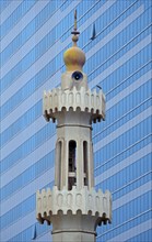 The minaret of a small mosque against the modern glass and steel facade of a skyscraper