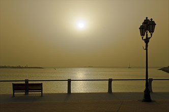 Sun setting over the Persian Gulf behind a bench and a street lamp at the Corniche