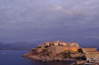 The citadel (castle) of Calvi surrounded by the mediterranean sea in front of the steep mountains of Corsica at the blue hour after sunset. Calvi is in the department Haute-Corse