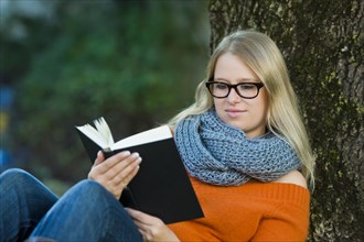 Young woman wearing glasses sitting in the park and reading a book