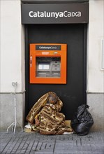 Homeless man sitting wrapped in a blanket in front of an ATM of the Spanish bank CatalunyaCaixa