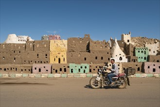 Man on a motorcycle passing by the mud-brick model of the old town of Bawiti