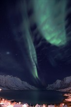 Aurora over a fjord with snow-covered mountains and an illuminated village