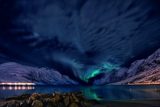 Aurora over a fjord with snow-covered mountains and an illuminated village