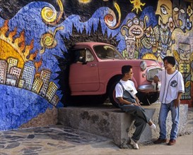 Young men talking in front of a sculpture of a car breaking through a wall