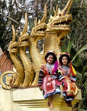 Two Lisu hill tribe girls wearing traditional costumes and headdresses sitting on a Naga sculpture