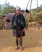 Elderly woman with a whistle standing in front of bamboo huts in an Akha village