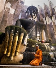 Praying monk neeling in front of the hand of the Buddha statue Phra Achana which is decorated with gold leaf