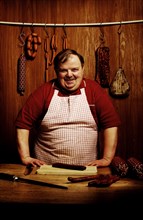 Fat butcher smiling with his hands propped on a bench in a butcher's store with a wooden ambience