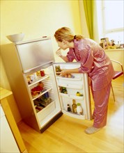 Woman wearing pyjamas standing at an open refrigerator and eating pickles