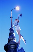 Woman wearing a summer dress standing on street lamp and holding hershoes in the air