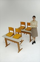 Strict female teacher standing next to old school desks with orange chairs