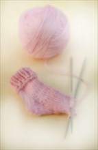 Semi-finished knitted pink baby sock with a ball of wool and knitting needles