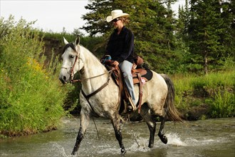 Cowgirl riding a gray horse through a river with splashing water