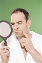 Man wearing a bathrobe observing himself critically in a hand mirror
