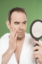 Man with eye cream observing himself critically in a hand mirror