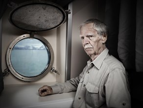 Elderly man stands in ship's cabin with a thoughtful expression